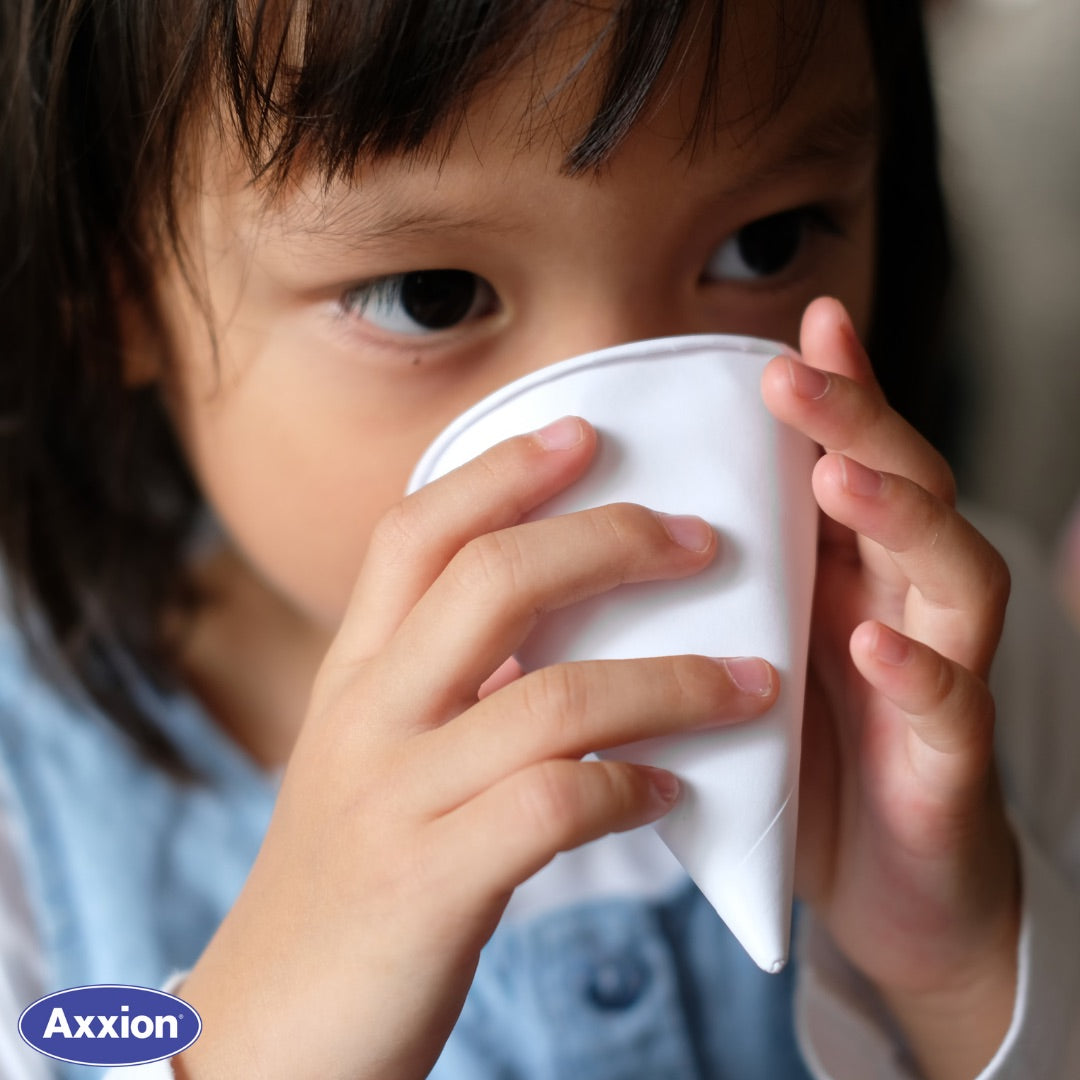 A close-up lifestyle image of a young child safely drinking from an Axxion 4.5 oz disposable white paper cone cup. Demonstrating the easy-to-hold, single-use design that is perfect for water coolers in schools, daycares, pediatric waiting rooms, and family-friendly facilities. The Axxion logo is visible in the bottom left corner.