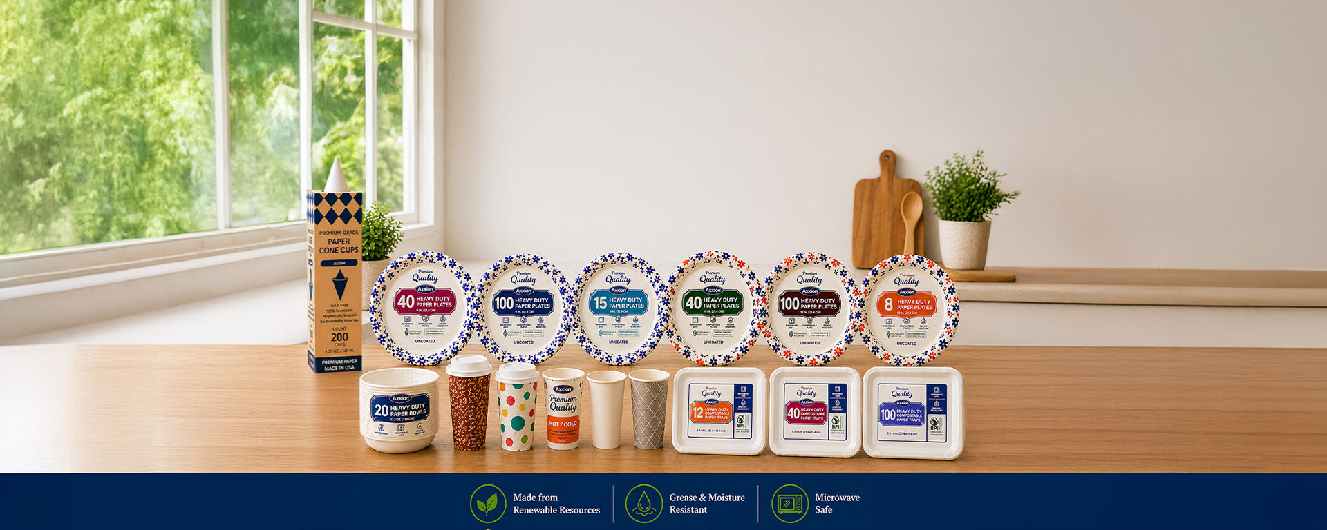 A wide assortment of Axxion premium heavy-duty paper plates, patterned cups, compostable paper cones and white food trays displayed on a wooden table in a bright, modern kitchen setting with a window. These disposable paper products are ideal for wholesale food service, catering, and restaurant supplies.