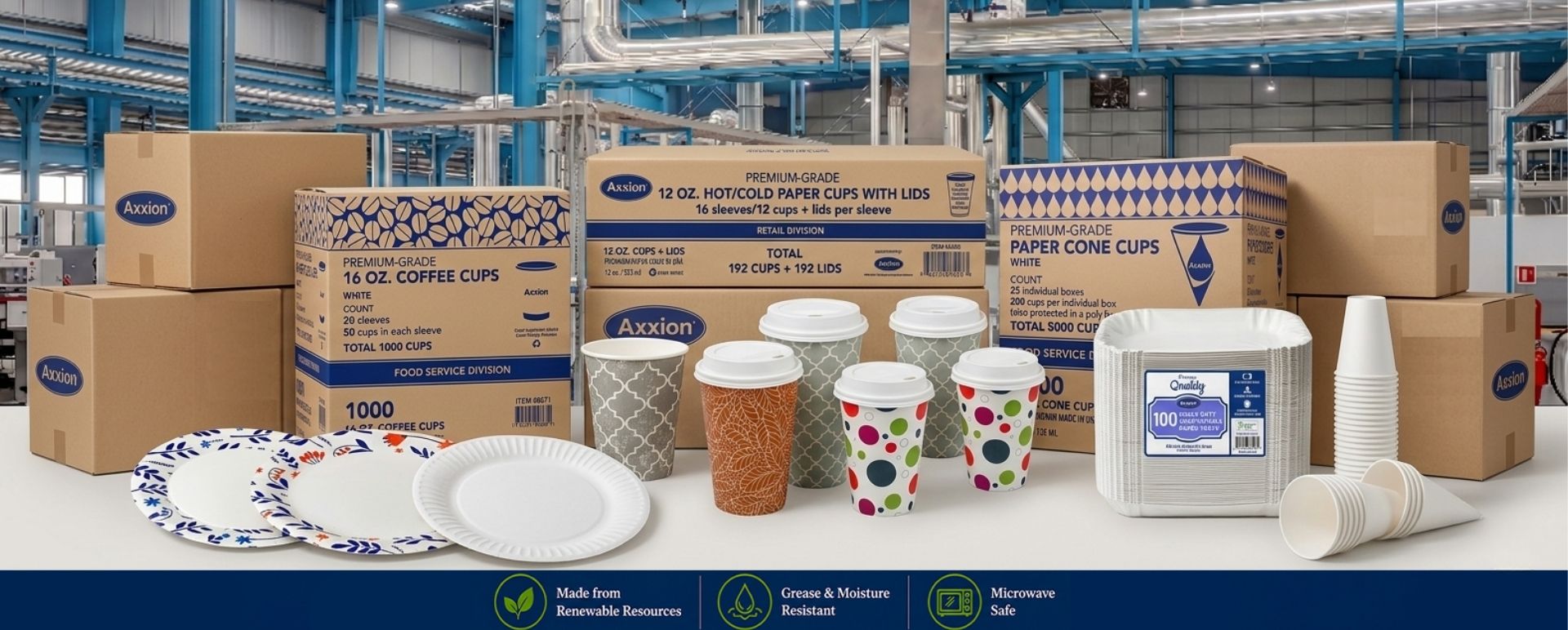 A comprehensive display of Axxion bulk disposable paper products arranged on a table inside a large industrial manufacturing facility. The collection features wholesale corrugated shipping boxes, patterned coffee cups with lids, white paper cone cups, heavy-duty white food trays, and designed and uncoated premium paper plates. Factory-direct paper goods ideal for restaurant supplies, commercial catering, and food service distributors.